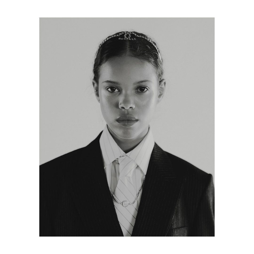 Portrait of a young woman with light brown skin, wearing a black blazer, white shirt, tie, necklace, and a jeweled headband.