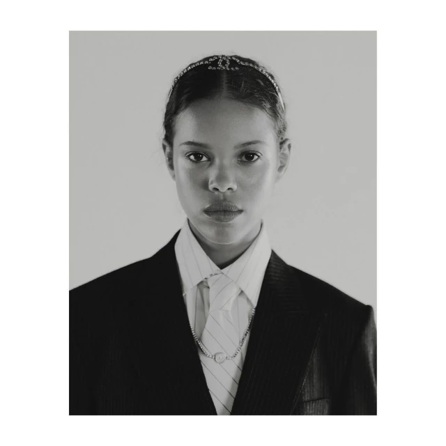 Portrait of a young woman with light brown skin, wearing a black blazer, white shirt, tie, necklace, and a jeweled headband.