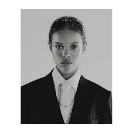Portrait of a young woman with light brown skin, wearing a black blazer, white shirt, tie, necklace, and a jeweled headband.