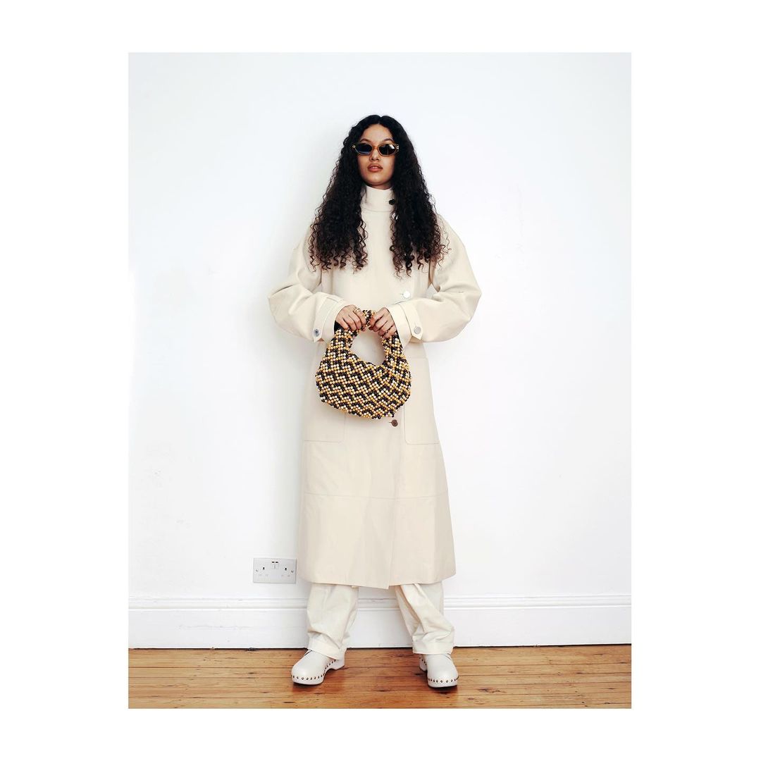 Full-length shot of a woman in a cream coat and pants, holding a small patterned handbag against a white wall and wood floor.