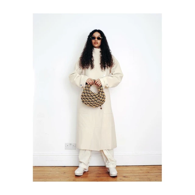 Full-length shot of a woman in a cream coat and pants, holding a small patterned handbag against a white wall and wood floor.