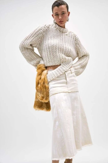 Woman in knit sweater and white skirt with faux fur stole. Fashion model poses in studio with a neutral background for a clothing advertisement.