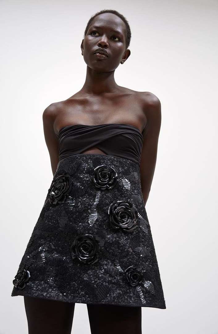 A Black woman models a black strapless top with a black floral embellished A-line skirt against a white background in a studio shot.