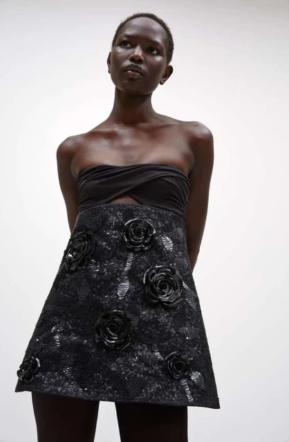 A Black woman models a black strapless top with a black floral embellished A-line skirt against a white background in a studio shot.