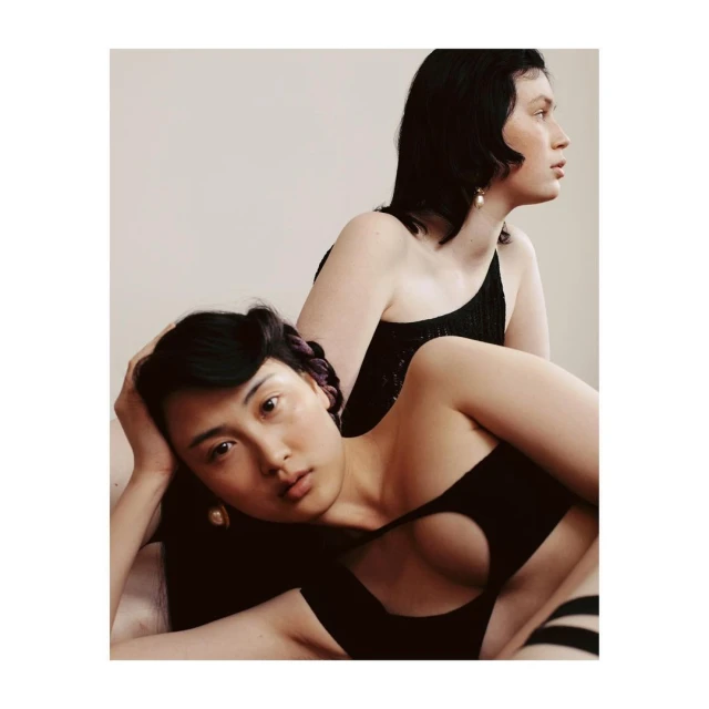 Two women pose in black dresses against a white background. One woman lies down looking at the camera while the other stands behind her.