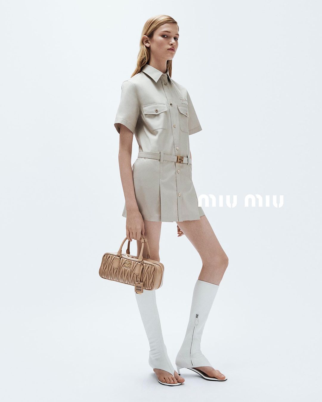 Blonde model wears a tan shirt dress, knee-high white boots, and holds a ruched bag in a studio with white background, fashion shot.