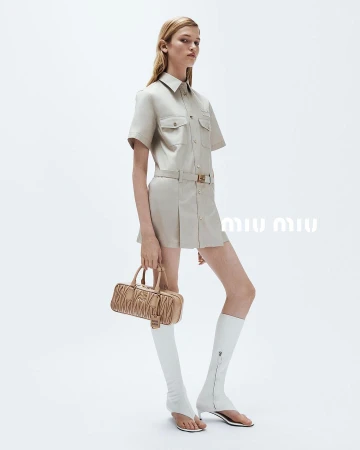 Blonde model wears a tan shirt dress, knee-high white boots, and holds a ruched bag in a studio with white background, fashion shot.