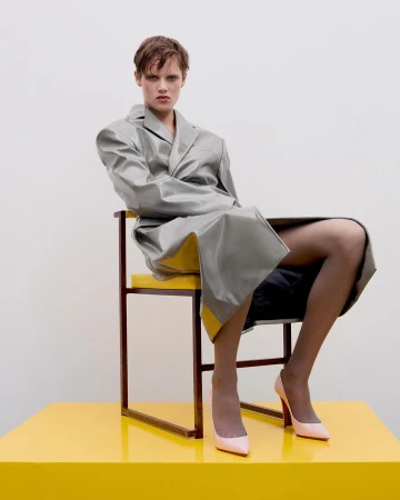 Fashion shot of a model in a grey coat and pink high heels sitting on a yellow chair against a white background in a photo studio.