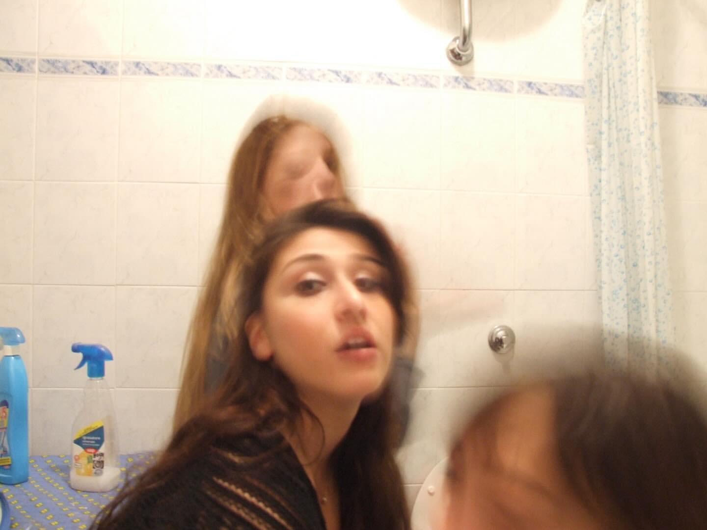 Close-up shot of two women in a bathroom with blue and white tiled walls and a shower curtain. The focus is soft due to motion blur.