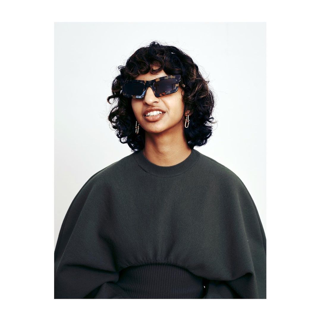 A woman with curly brown hair and sunglasses poses indoors, wearing a dark green top against a white background in a medium shot.