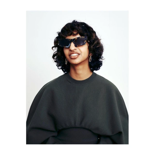 A woman with curly brown hair and sunglasses poses indoors, wearing a dark green top against a white background in a medium shot.