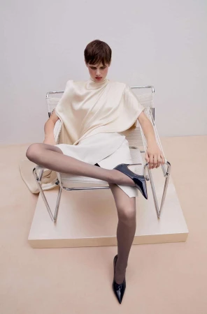 Model with short hair seated in a chrome armchair wearing cream top, white skirt, sheer grey tights, and black high heels