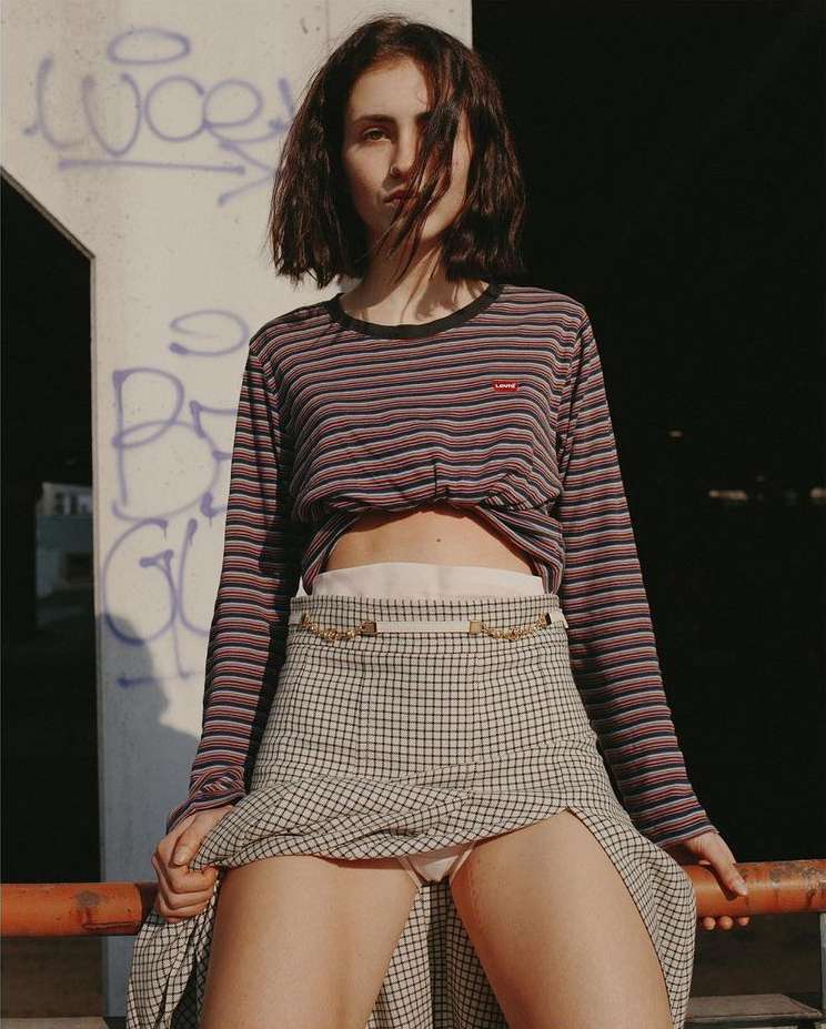 Young woman poses outdoors with a plaid skirt and long-sleeved top, sitting on a railing with graffiti in the background.