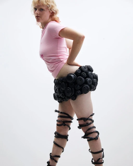 Adult female fashion model in a light pink t-shirt, black skirt, and black straps around the legs posing in a bright white studio.