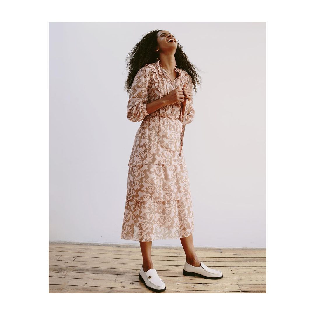 Full shot of a curly-haired woman in a peach-colored, floral-print dress and white loafers laughing against a white backdrop.