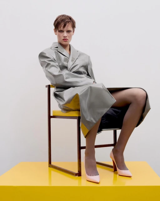 A fair-skinned adult female model in a grey trench coat sits on a yellow and brown chair against a pale grey background in studio lighting.