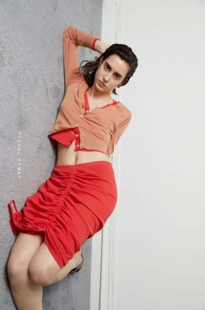 Woman in orange cardigan and red skirt reclining on grey carpeted floor by a white wall