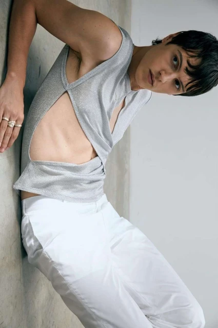 Fashion shot of a slender adult man in a metallic tank top and white pants leaning against a wall in a photo studio.