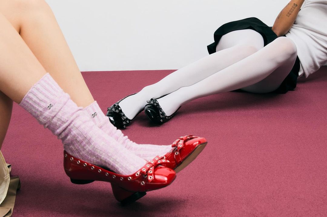 Close-up of two women lying on a mauve surface, legs crossed. One wears red ballet flats and pink socks, the other black ballet flats and white tights.