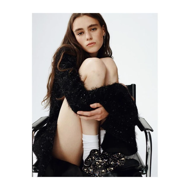 Adult female posing in a black cardigan while sitting on a black chair in front of a white background in a studio environment.