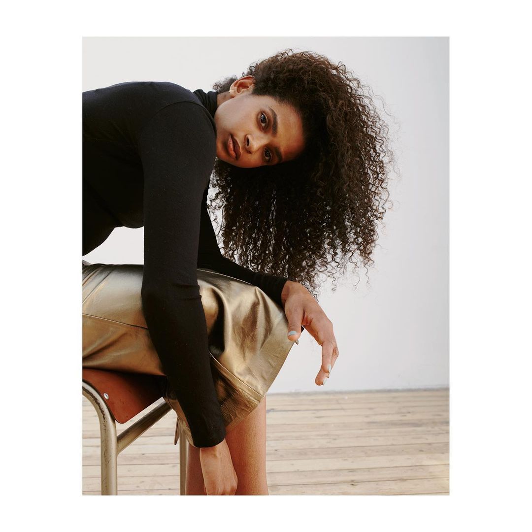 Woman with curly hair in black top poses leaning on chair with metallic gold skirt in a minimalist studio with wood floor.