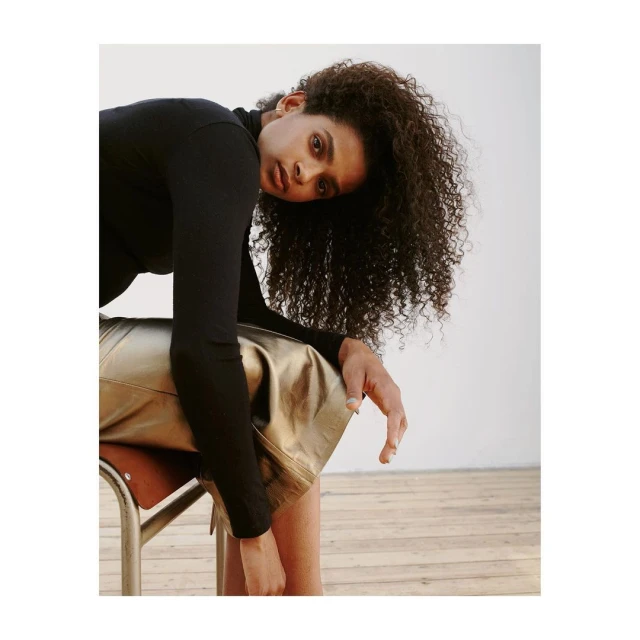 Woman with curly hair in black top poses leaning on chair with metallic gold skirt in a minimalist studio with wood floor.