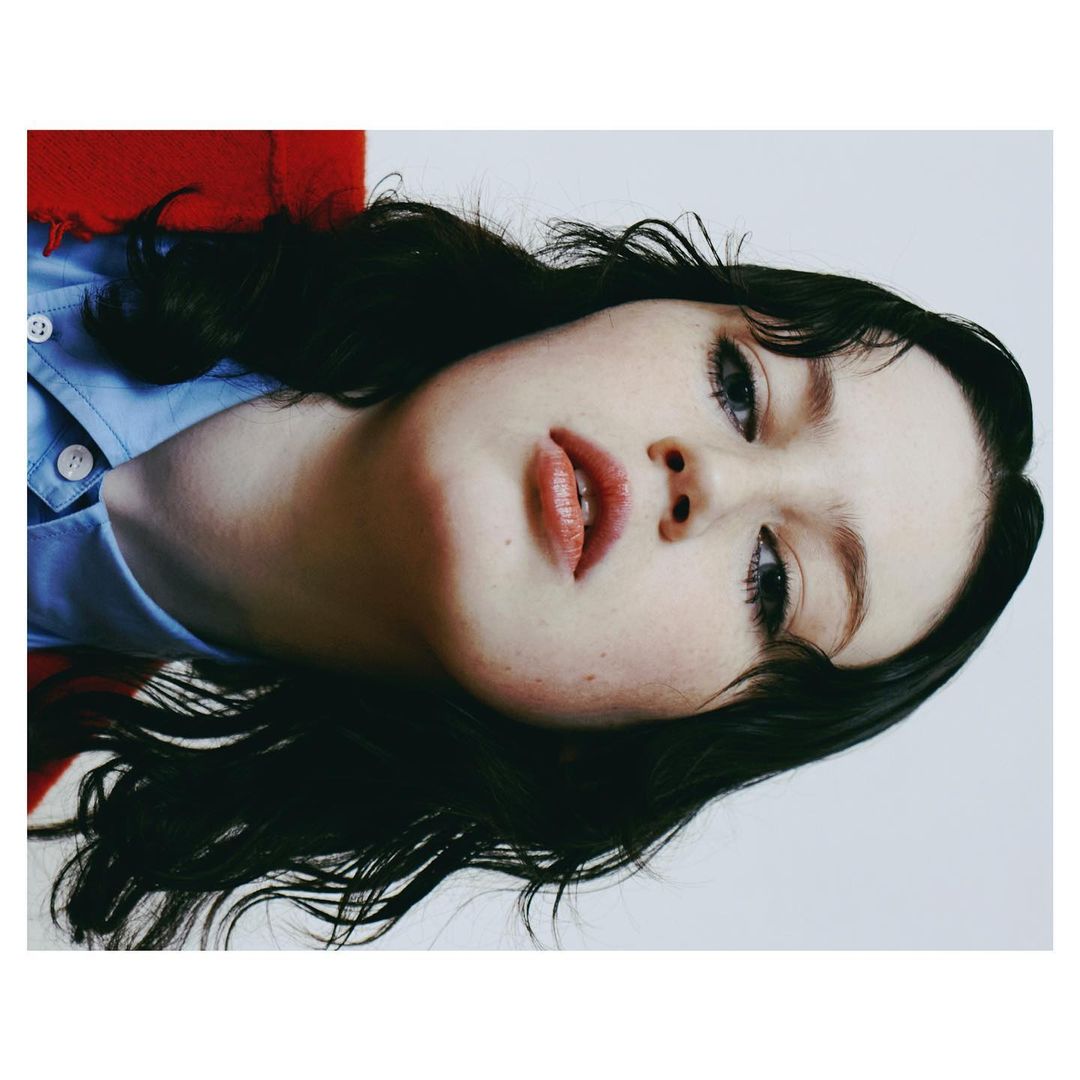 Close-up portrait of a young adult woman with dark wavy hair, wearing a blue shirt, set against a white background with red fabric.