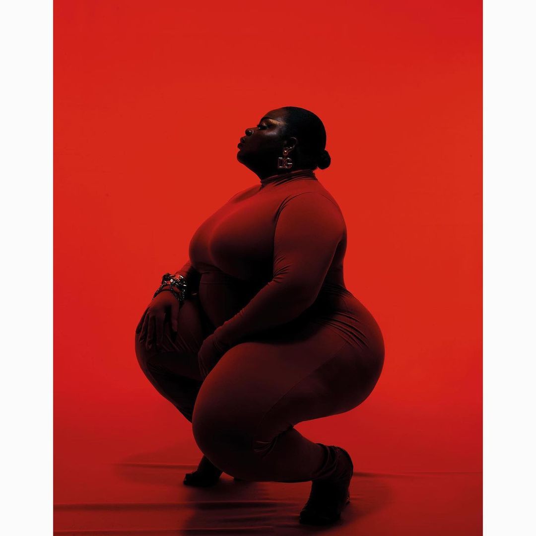 Portrait of a curvy black woman in a matching red outfit, posed in a squat against a solid red backdrop, emphasizing body positivity.