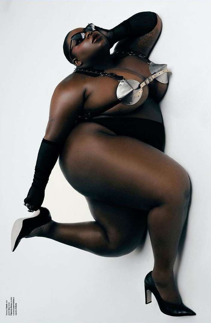 Adult female with an athletic build poses on a white surface with a metallic bra top, black sunglasses, gloves, high heels, and dark undergarments.