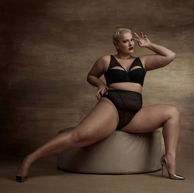A curvy woman with short blonde hair poses in a black lingerie set and metallic silver high heels in a studio setting with neutral backdrop.