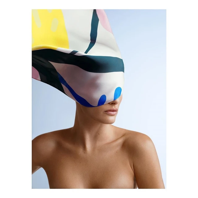 A woman with bare shoulders poses in front of a light blue background, wearing a colorful patterned silk scarf over her head.