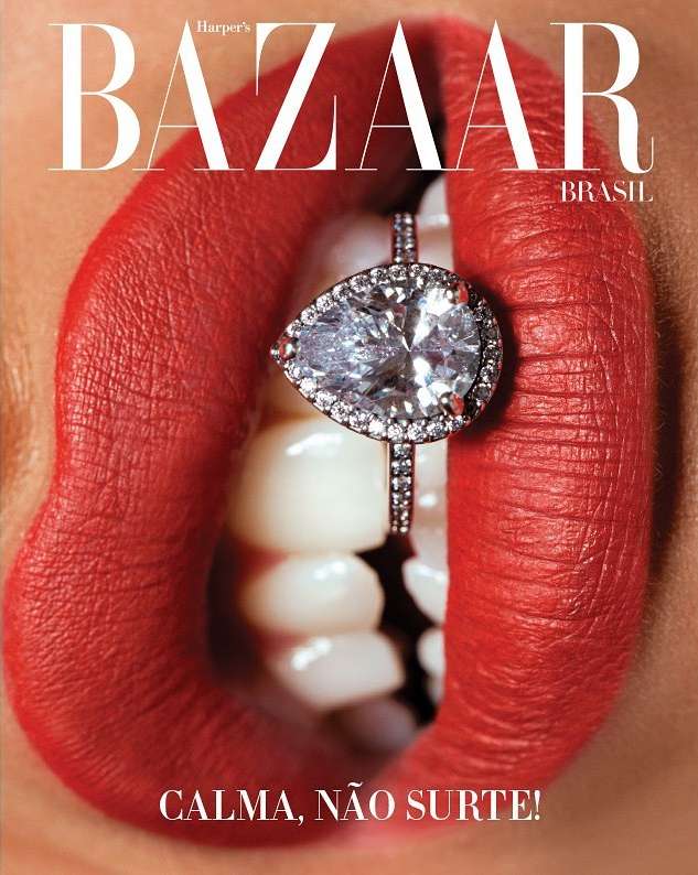 Close-up of a woman's red lipstick-covered lips holding a diamond ring between her teeth against a magazine cover backdrop.
