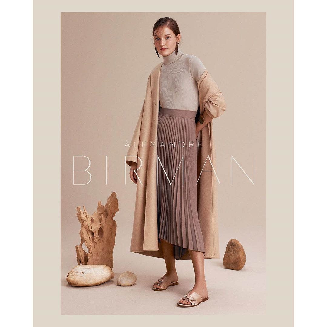Elegant woman in a tan turtleneck, pleated skirt, and coat, posing in a studio with natural stone elements, creating a fashion editorial.