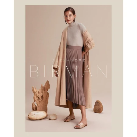 Elegant woman in a tan turtleneck, pleated skirt, and coat, posing in a studio with natural stone elements, creating a fashion editorial.
