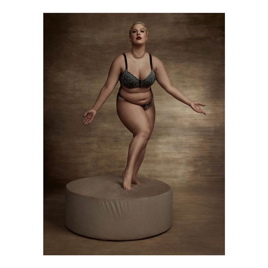 Full-figured female model in lingerie stands on a round platform in a photo studio with a brown mottled backdrop and soft lighting.