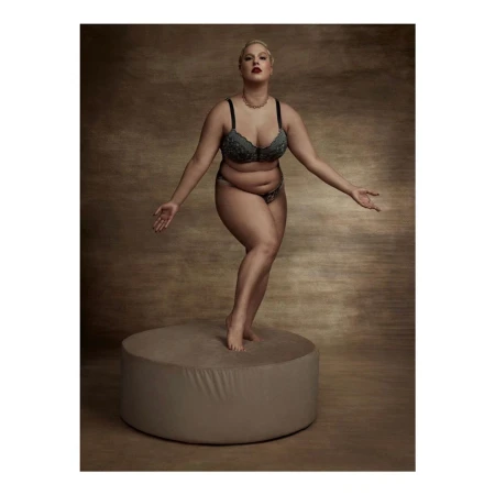 Plus size woman in matching bra and briefs balancing on a circular pedestal against a mottled dark background.