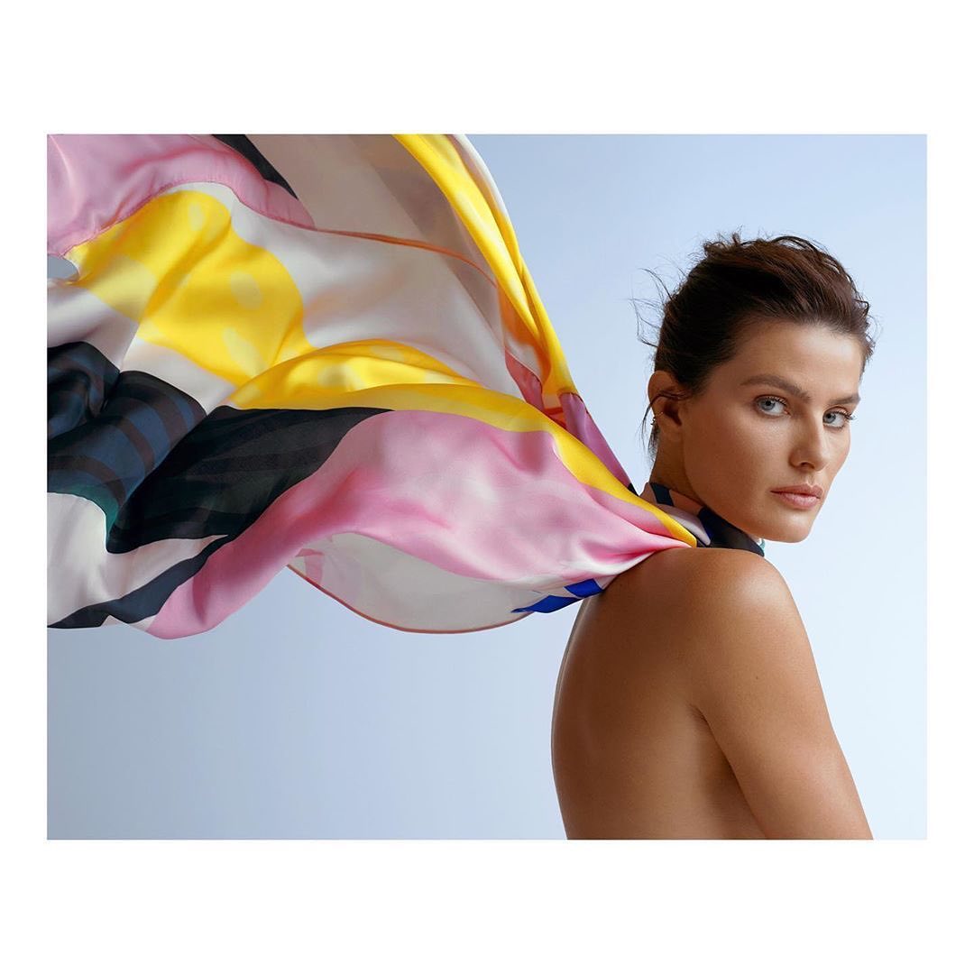 Portrait of a brunette woman with a colorful abstract-print scarf draped over her shoulder, set against a soft, graduated blue backdrop.