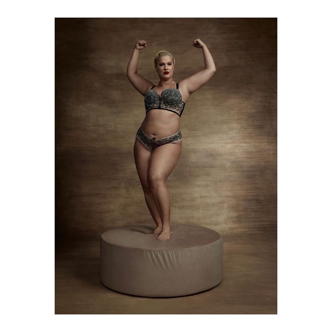 Blonde woman in lingerie poses on a stand in a studio with arms raised, showcasing her figure and physique in a portrait.