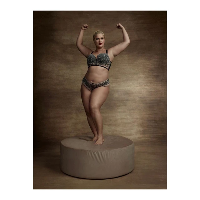 Blonde woman in lingerie poses on a stand in a studio with arms raised, showcasing her figure and physique in a portrait.