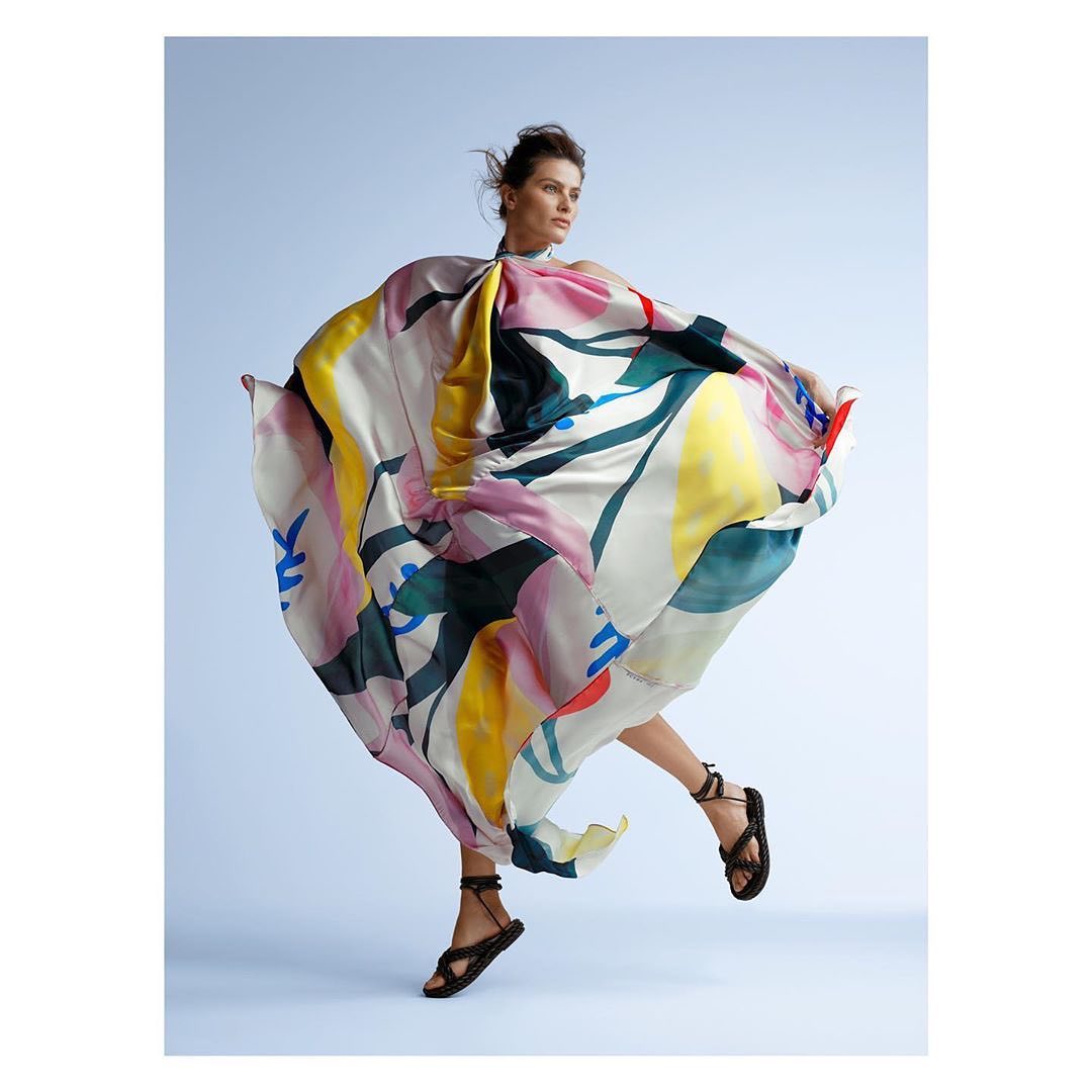 Woman in a vibrant, flowing dress leaps gracefully in a light blue studio, showcasing fashion and style with a blend of art and motion.