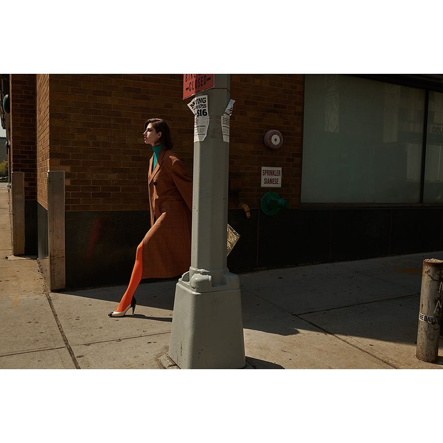 A woman in a brown coat walks on a sidewalk in an urban setting wearing orange stockings, green turtleneck, and white high heels.
