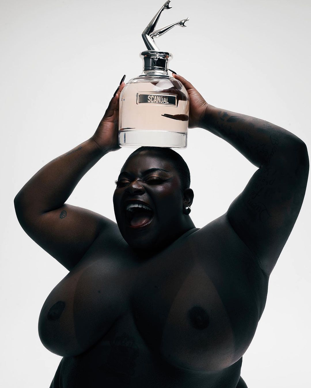 An adult Black woman holds a bottle of Scandal perfume overhead against a stark white background, with a joyful facial expression.