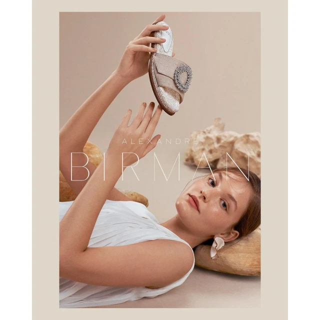 Fashion shot featuring an adult female model lying down holding a light brown slide sandal with a jeweled embellishment and a beige background.