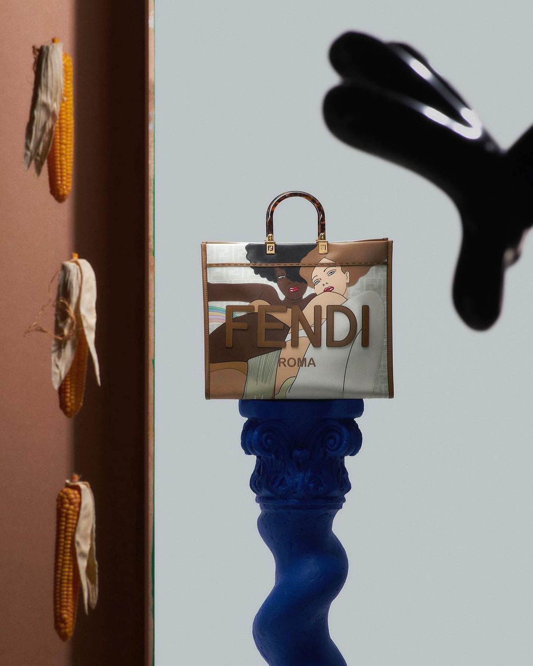 Fendi handbag displayed on a blue pillar, featuring an illustration, against a light blue background with corn cobs and a black object.