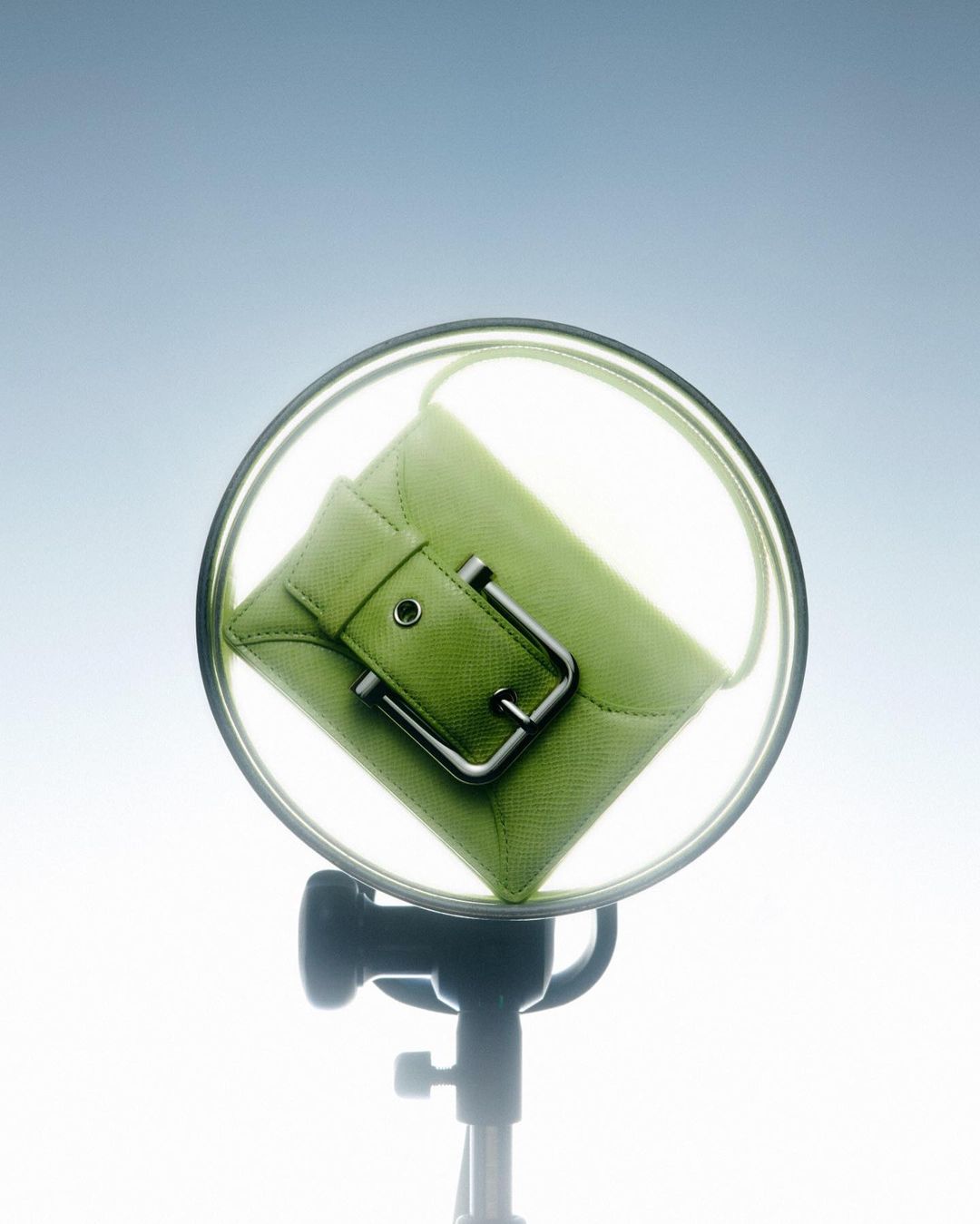 A light green leather clutch with a buckle is displayed in front of a studio light on a light blue gradient background.