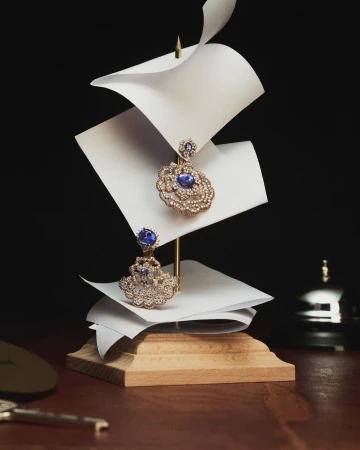 Elegant jewelry display featuring gold earrings with a central blue stone, balanced on white paper sheets, against a dark background.