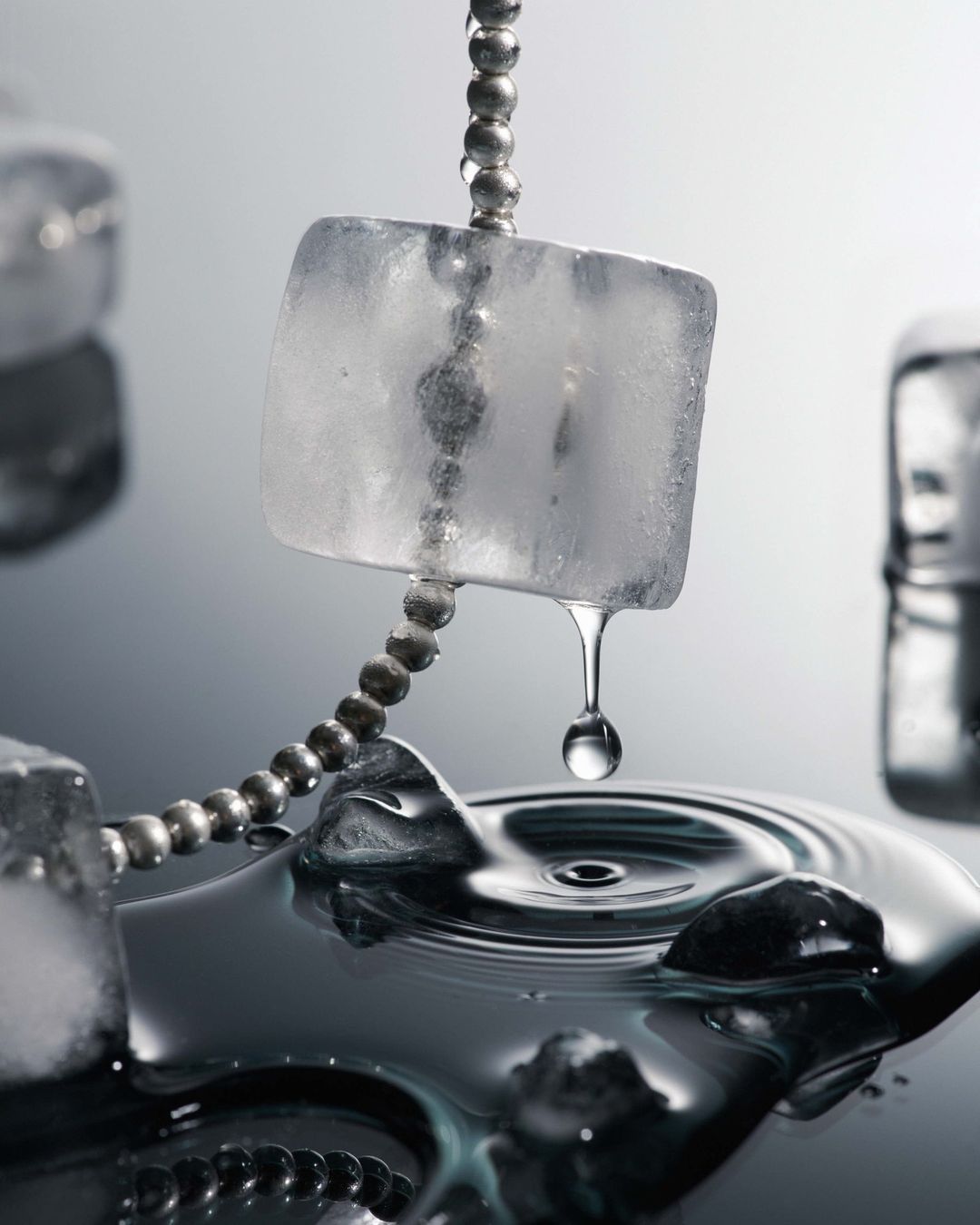 Close-up of ice cubes suspended on a beaded chain with a water droplet falling into a pool creating ripples on a reflective surface.