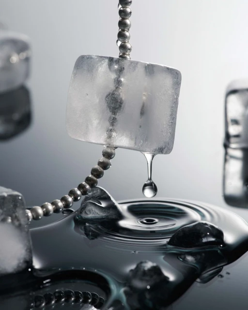 Close-up of ice cubes suspended on a beaded chain with a water droplet falling into a pool creating ripples on a reflective surface.