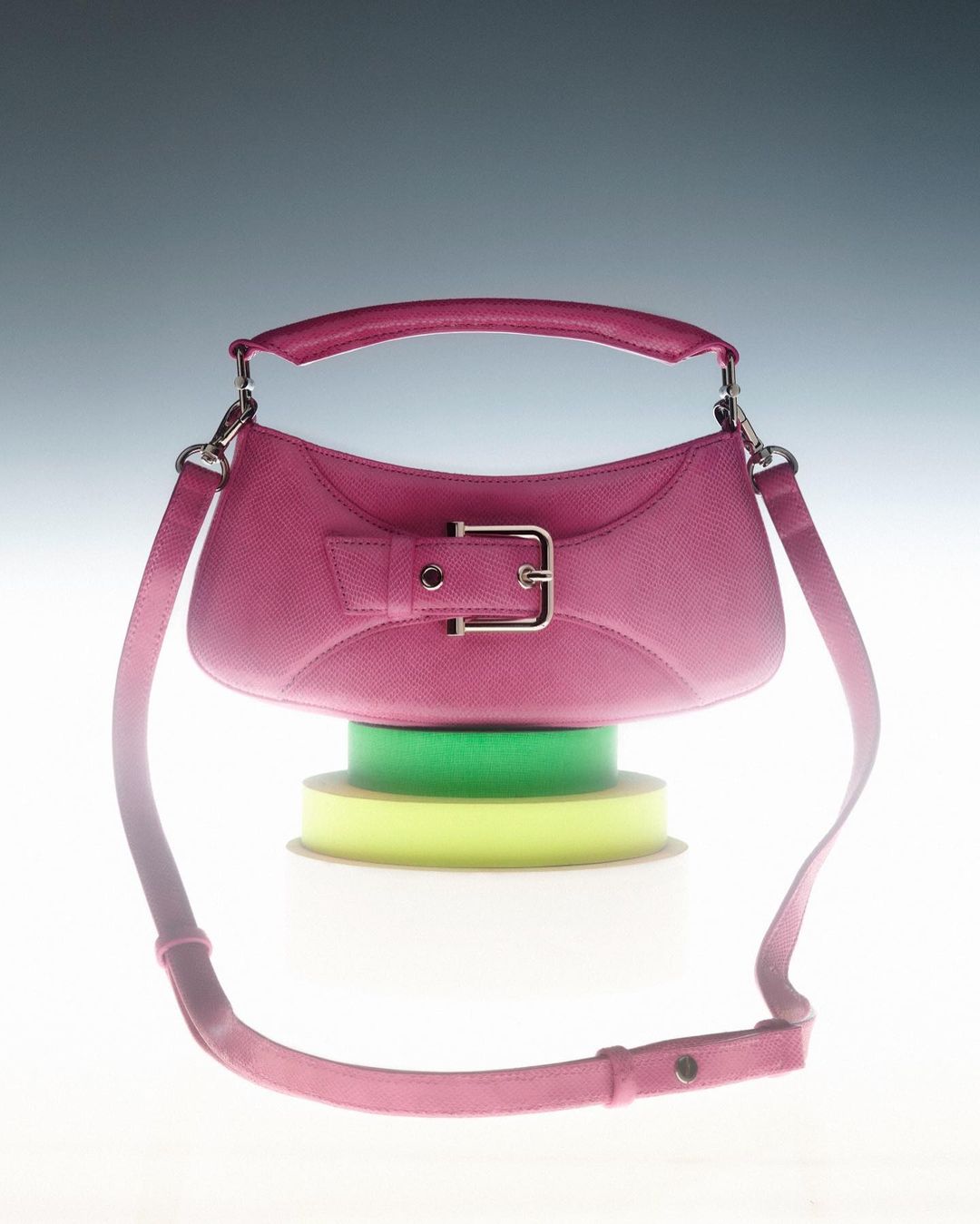Studio shot of a pink leather handbag with a silver buckle, resting on rolls of yellow and green ribbon against a grey background.