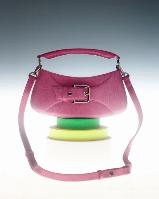 Studio shot of a pink leather handbag with a silver buckle, resting on rolls of yellow and green ribbon against a grey background.